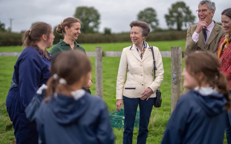 Princess Anne stands in a field talking with children in blue overalls and rain jackets