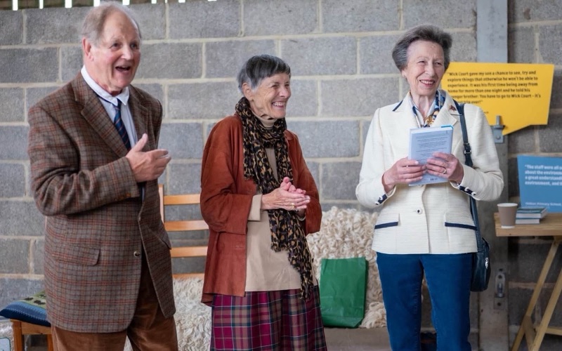 Princess Anne stands holding a booklet beside a man in a tweed jacket and a woman who is laughing