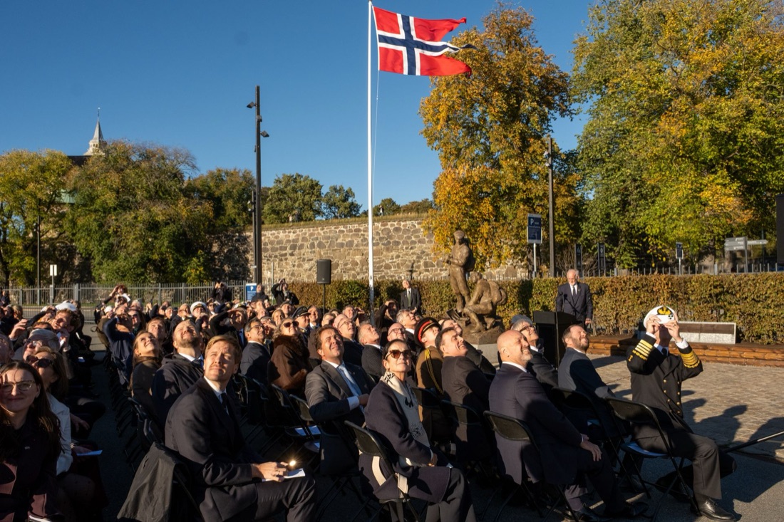 Publikum på Normandieplassen ser opp mot himmelen under Spitfire-overflyvningen, med det norske flagget og monumentet i bakgrunnen
