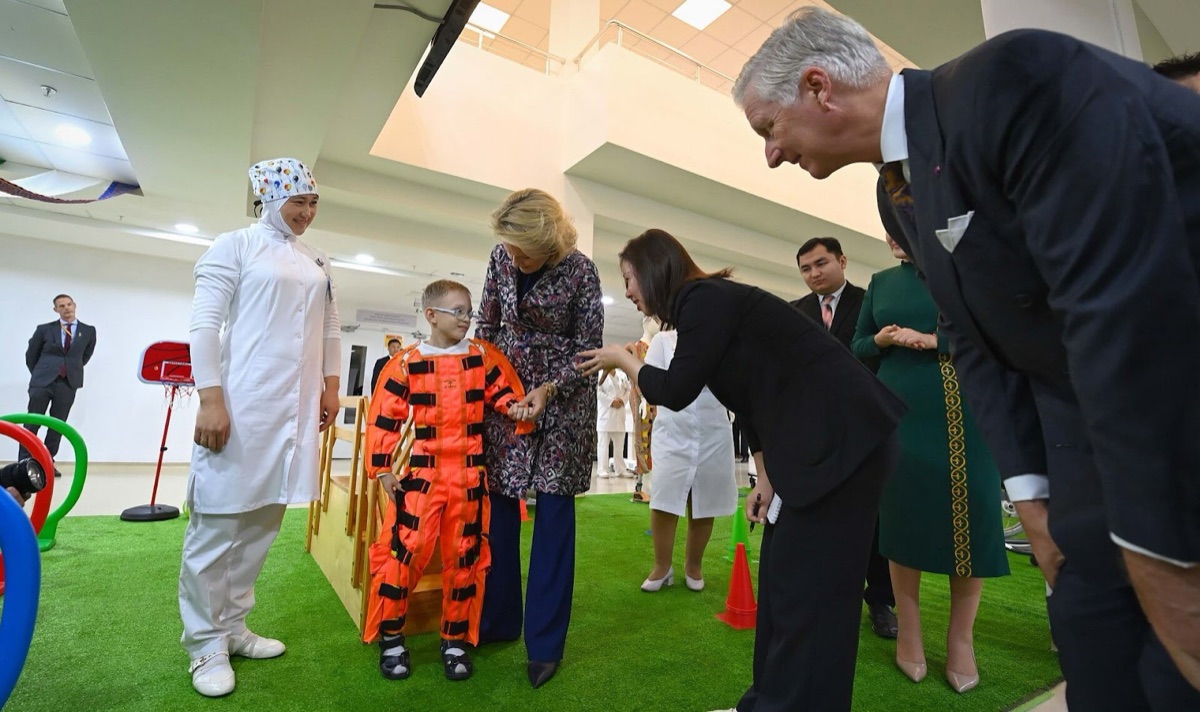 Queen Mathilde holds the hand of a boy in an orange training suit while King Philippe bends down beside them
