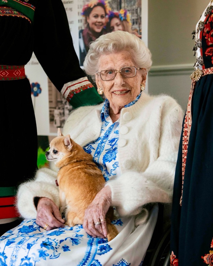 Princess Astrid sits smiling with her dog Wilma in her lap, surrounded by national costumes on display mannequins
