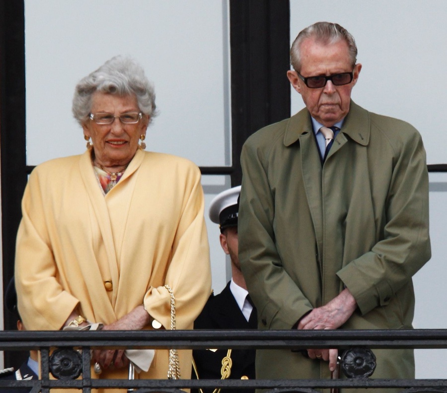 Princess Astrid in a yellow coat stands on a balcony beside an elderly man in a green coat and sunglasses