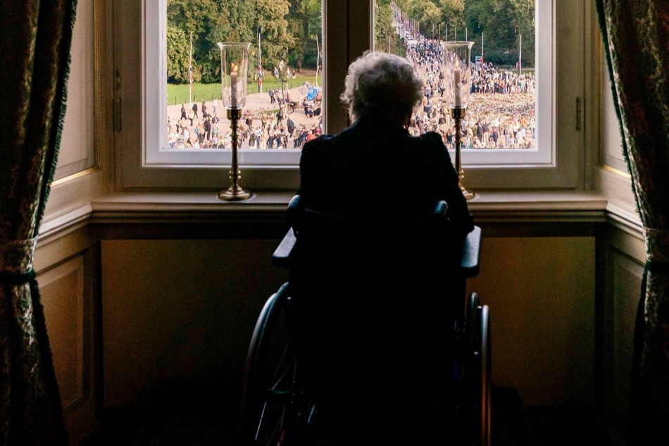 Princess Astrid seen from behind in a wheelchair at a Palace window, with two candles on the sill and the crowds outside