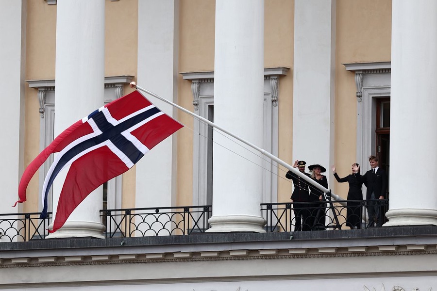 The Palace balcony seen from the side with the Norwegian flag flying at the top of the pole and the royal family standing on the balcony behind