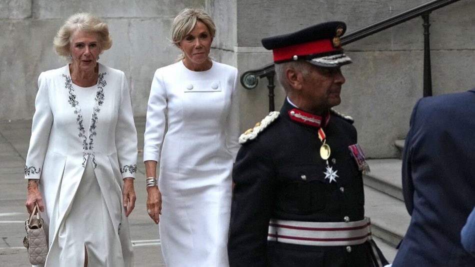 Queen Camilla and Brigitte Macron walk side by side, both in white, with a uniformed official in the foreground