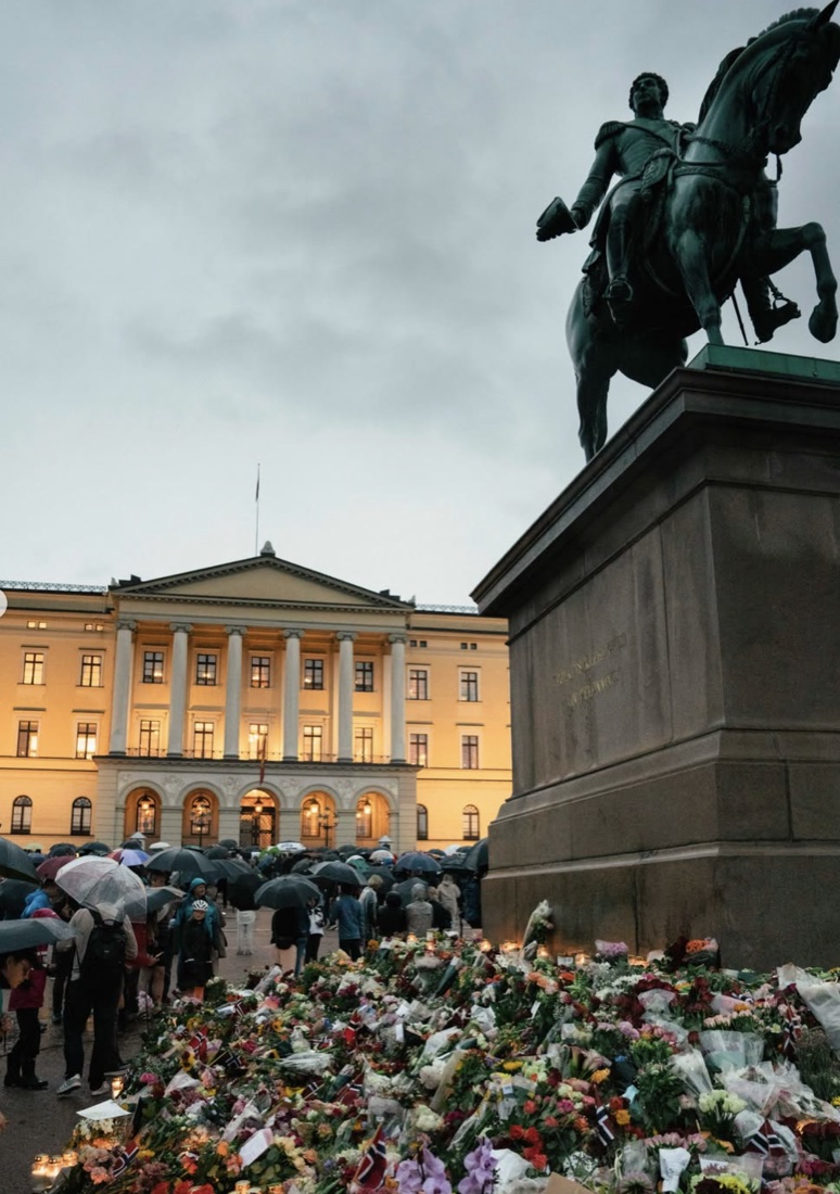 Slottsplassen i regnvær med paraplyer, blomsterhav foran statuen av Karl Johan og Slottet opplyst i bakgrunnen