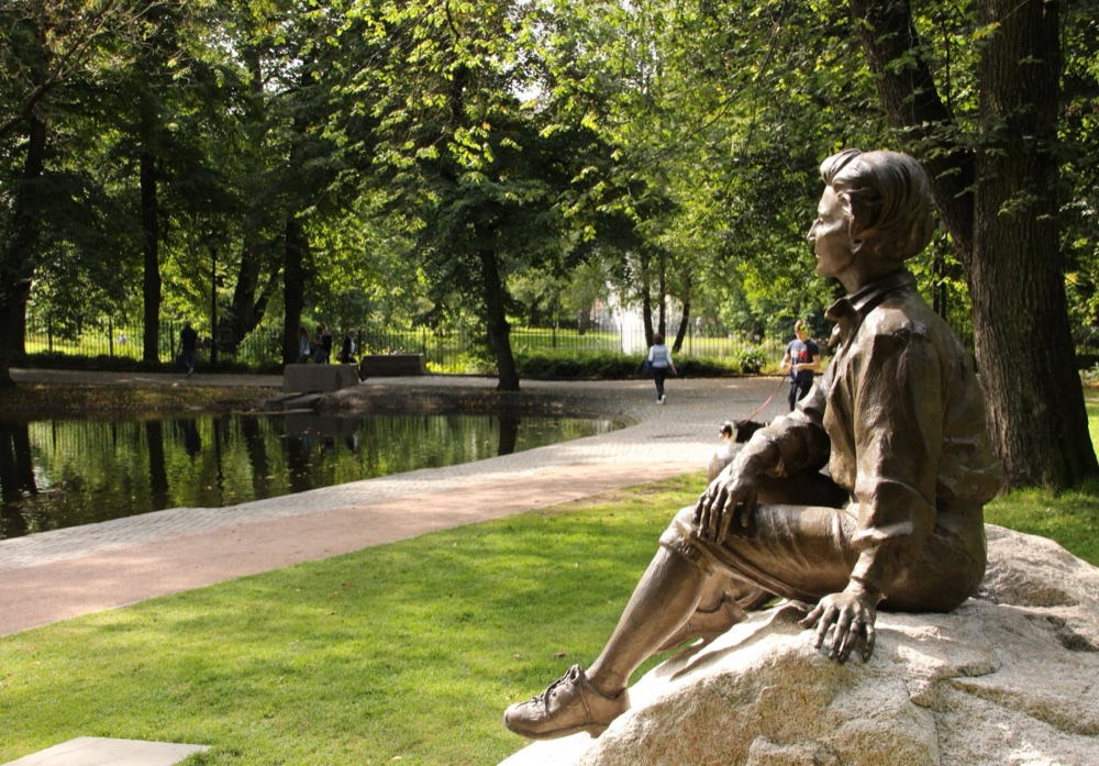 Bronsestatue av dronning Sonja sittende på en stein med tursekk, med utsikt over en park