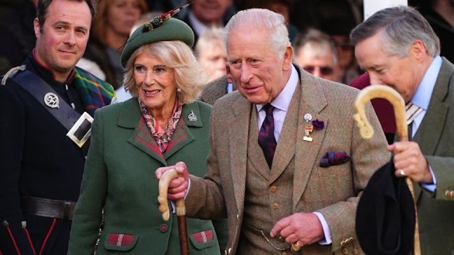 King Charles in a tweed jacket and Queen Camilla in a green coat and green hat walk side by side among people in Highland dress