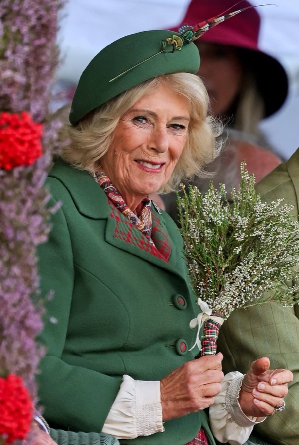 Close-up of Queen Camilla in a green coat and green hat with a pheasant feather, holding the heather bouquet