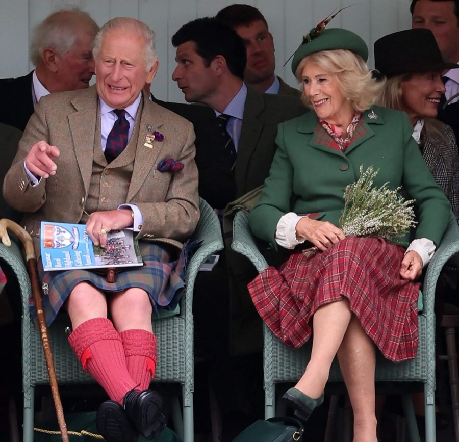 King Charles in a tweed jacket and kilt sits beside Queen Camilla in a green coat in the stand, holding the programme