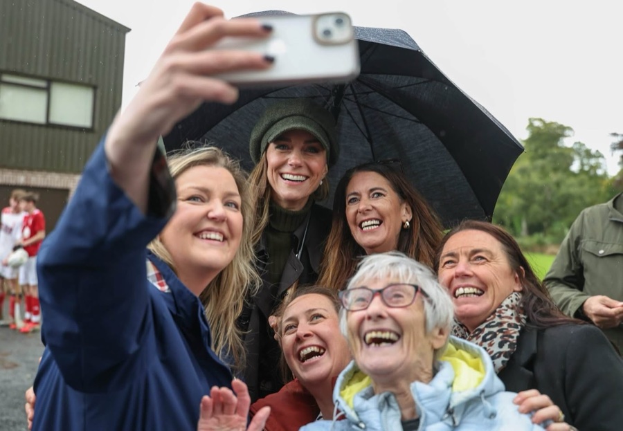 Princess Catherine smiles under an umbrella as five women take a selfie with her in the rain