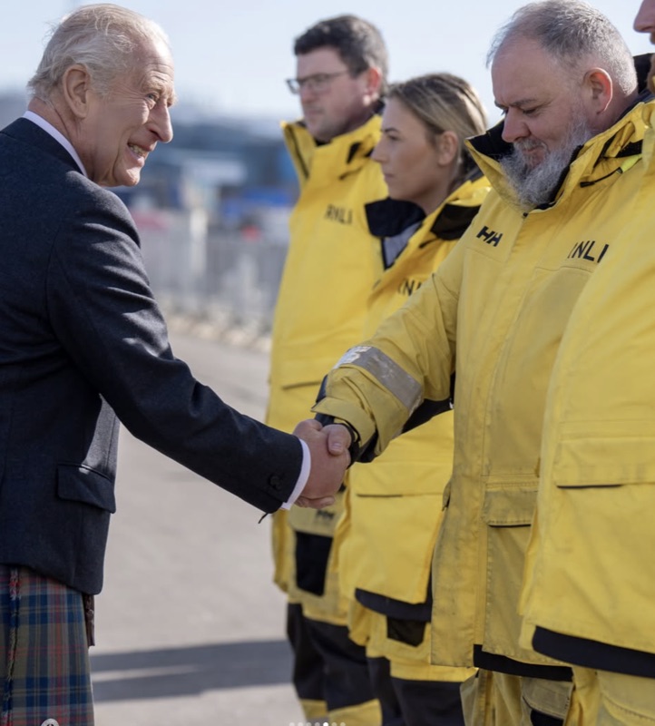Kong Charles håndhilser på en mann i gul redningsdrakt merket RNLI, med to andre mannskaper ved siden av