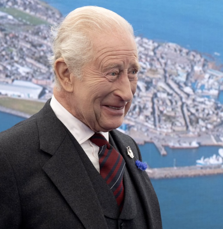 Portrett av kong Charles foran en storskjerm som viser et flyfoto av havn og bebyggelse