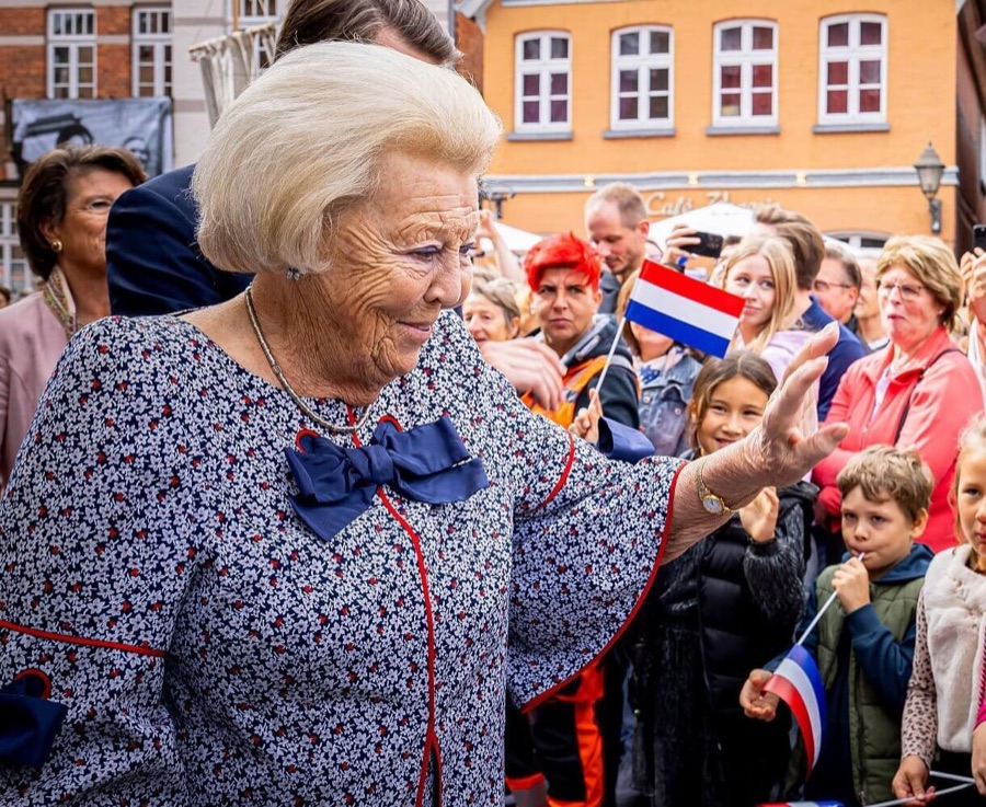 Princess Beatrix waves to onlookers holding Dutch flags in a street lined with red and yellow brick buildings