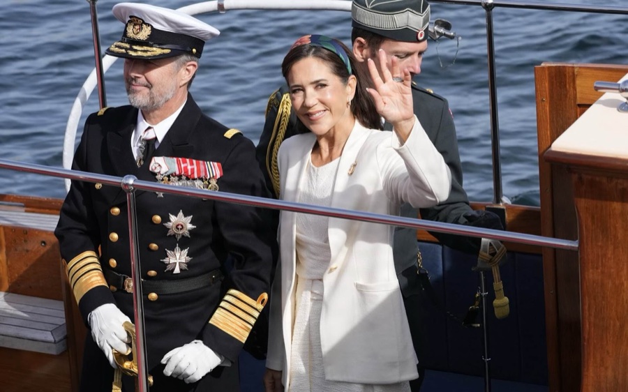 King Frederik in naval uniform with a white peaked cap stands on board the royal yacht while Queen Mary in white waves