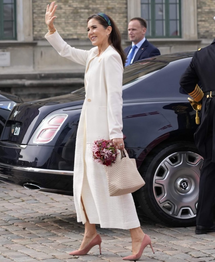 Queen Mary in a white coat waves with a bouquet in her hand beside a dark car on the cobblestones