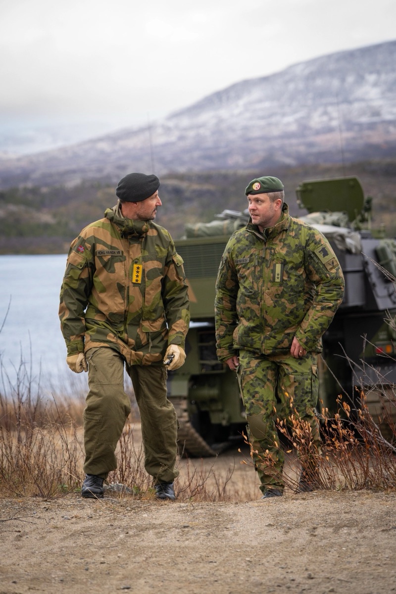 Kongen går sammen med en offiser langs et beltekjøretøy i terrenget