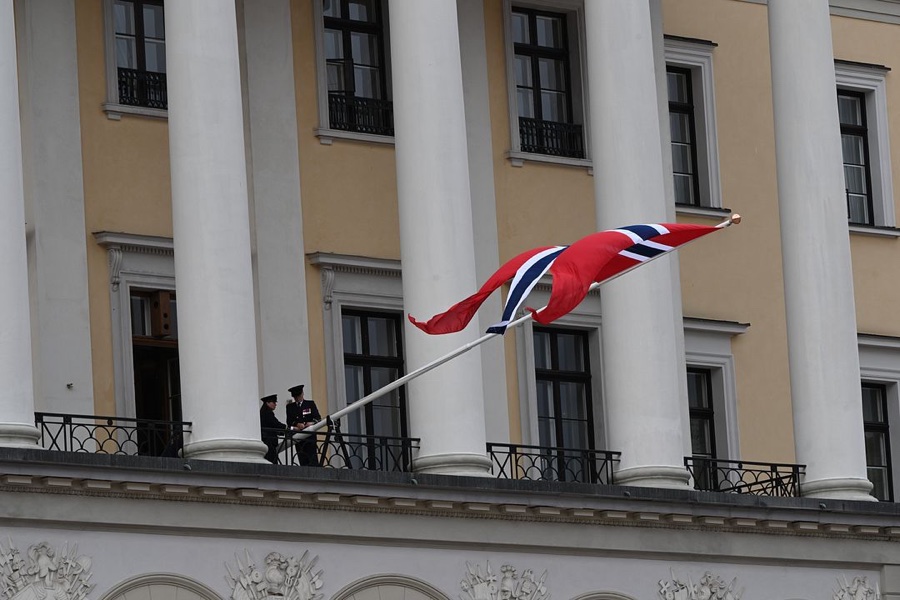 The Norwegian flag flies at the top of the flagpole at the Royal Palace, with two uniformed figures on the balcony behind
