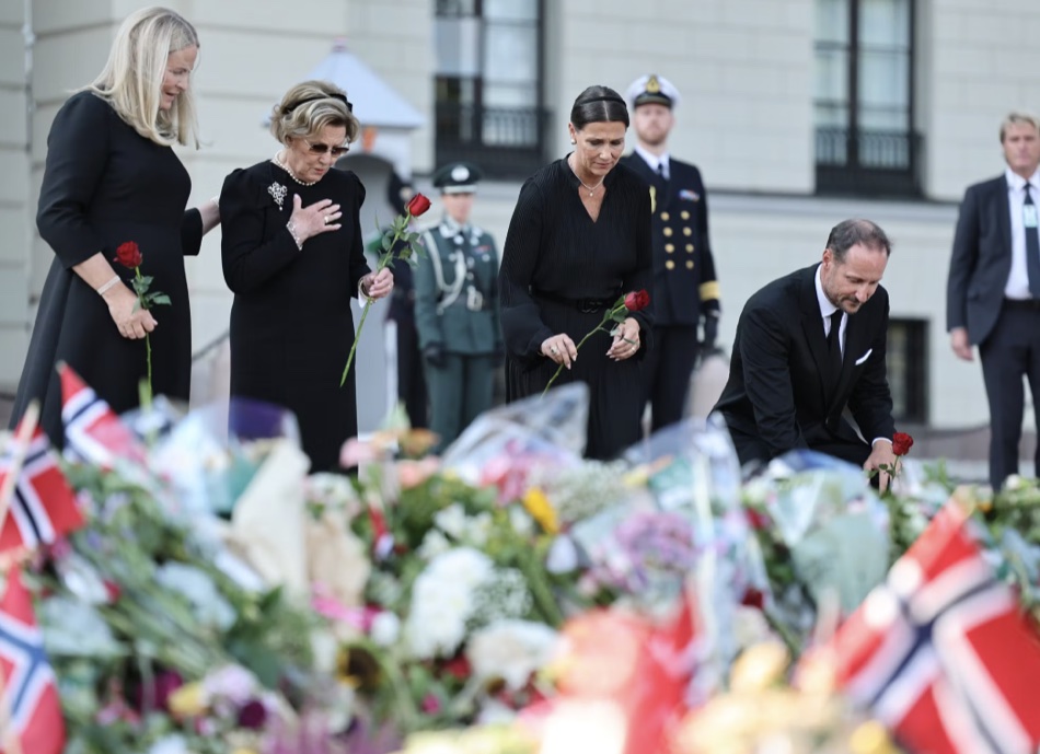 Dronning Mette-Marit, dronning Sonja og prinsesse Märtha Louise står med røde roser mens kong Haakon setter seg ned på huk for å legge sin rose i blomsterhavet