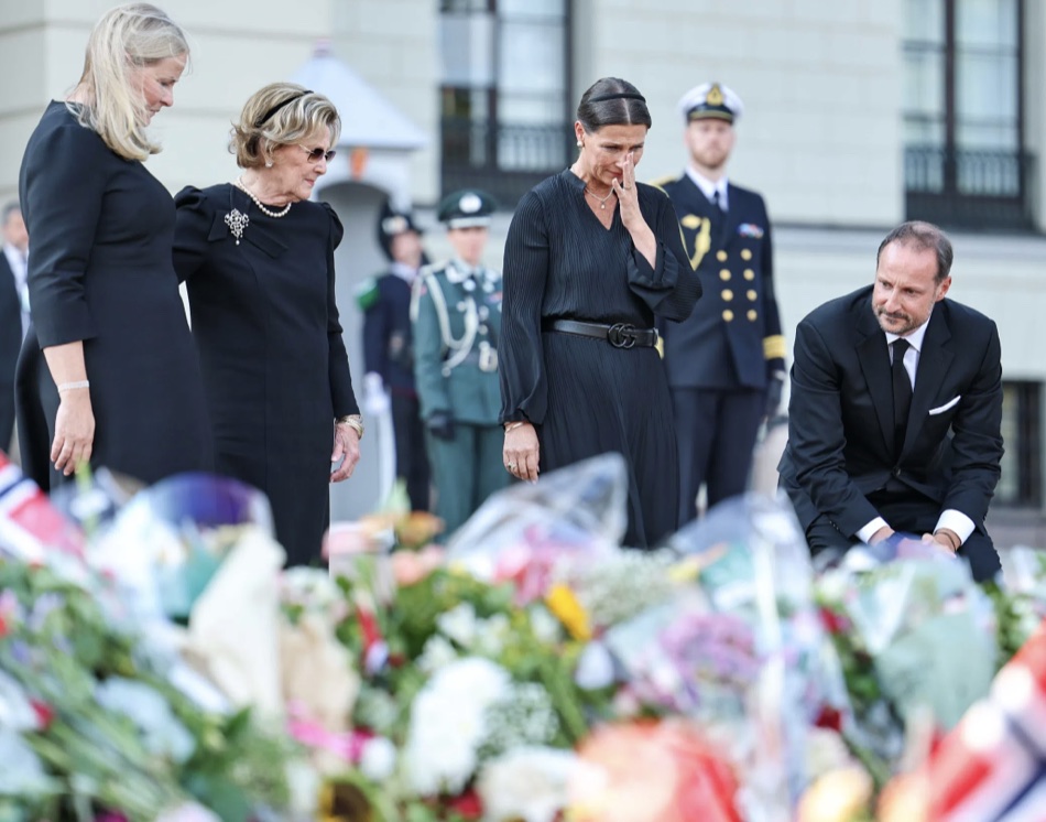 Dronning Mette-Marit, dronning Sonja og prinsesse Märtha Louise ser ned mot blomstene, prinsessen tørker tårer, mens kong Haakon sitter på huk ved blomsterhavet