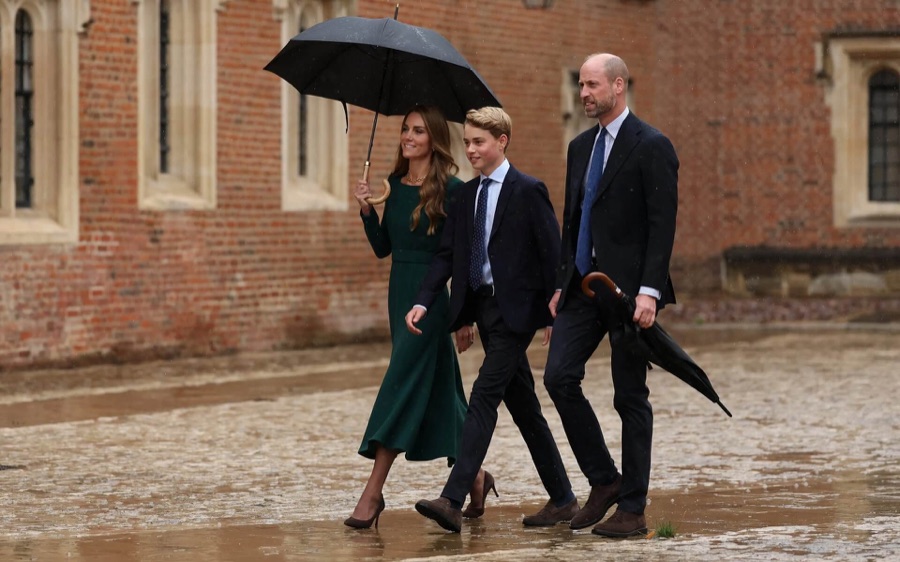 The family walk through the rain across a courtyard with red brick buildings behind them, the Princess with an umbrella and Prince George in the middle