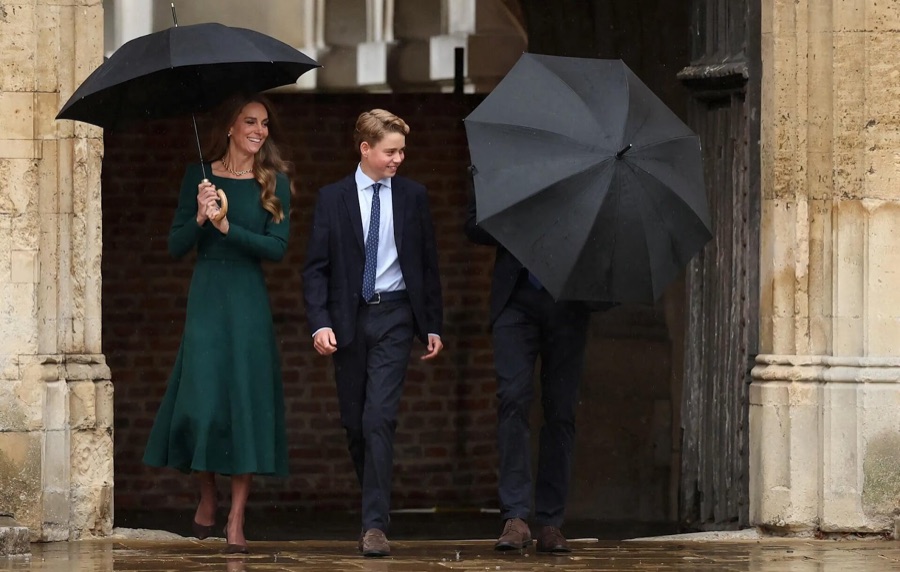 The Princess of Wales laughs as she walks under the umbrella beside Prince George in a dark suit, coming out through an archway
