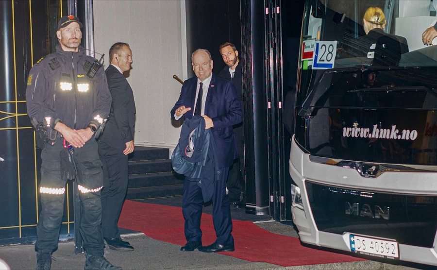 Prince Albert stands on a red carpet outside the Grand Hotel beside a coach, with an armed police officer on the left of the picture