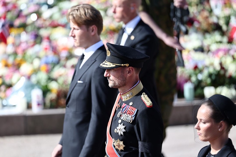 Close-up of King Haakon in full dress uniform with orders, in profile, with the sea of flowers blurred behind him
