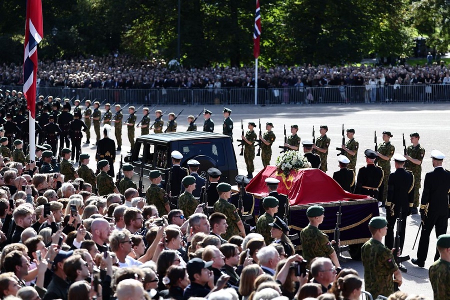 The coffin draped in the royal standard rests on a gun carriage with white flowers, surrounded by soldiers with rifles and dense crowds behind the barriers