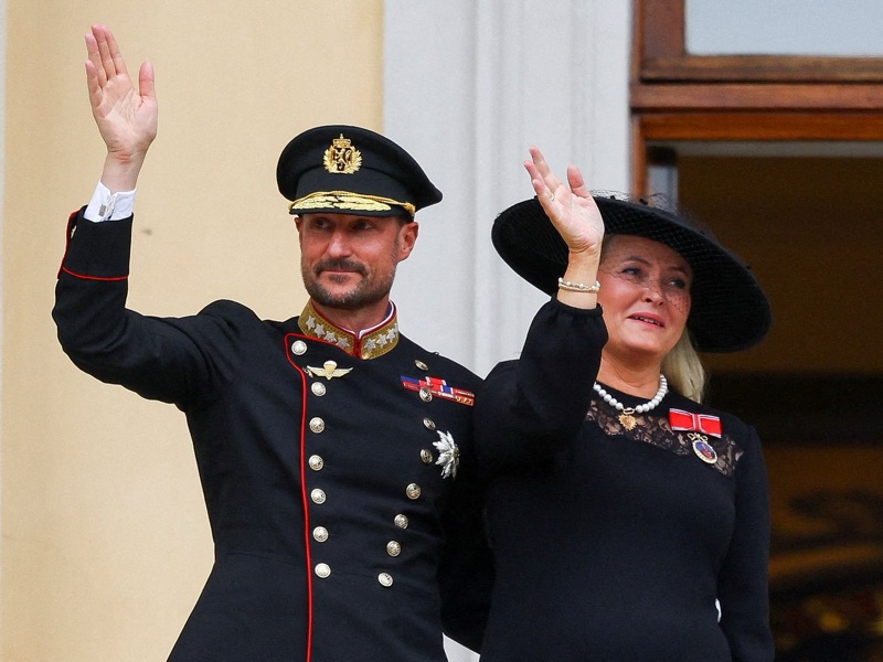 King Haakon in gala uniform and Queen Mette-Marit dressed in black waving from a balcony