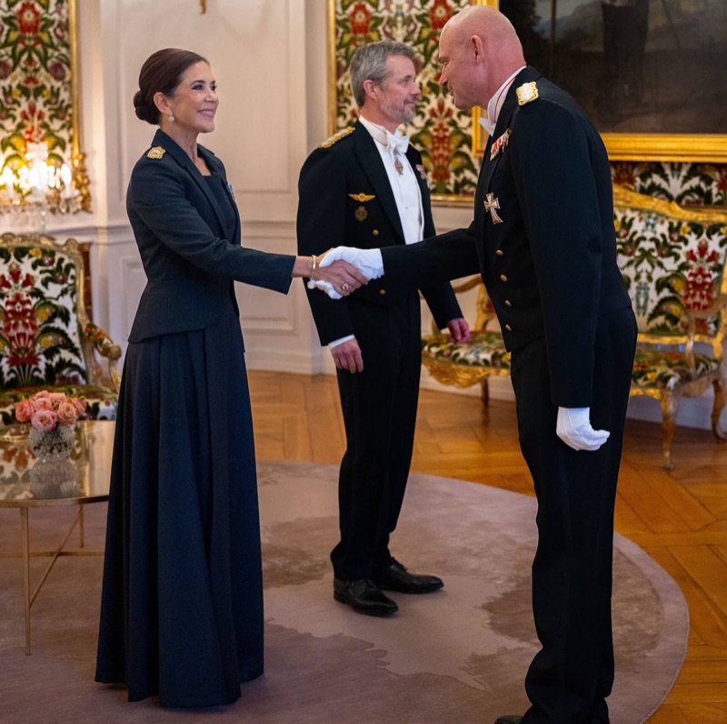Dronning Mary håndhilser på en offiser i uniform med hvite hansker, mens kong Frederik står bak