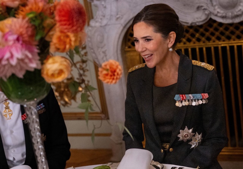 Dronning Mary sitter ved taffelet og smiler, med store blomsteroppsatser på bordet