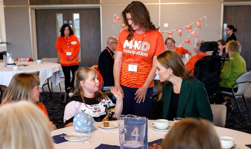 Prinsesse Catherine bøyer seg ned i samtale med en jente i rullestol ved et bord, med frivillige i oransje MND-trøyer rundt