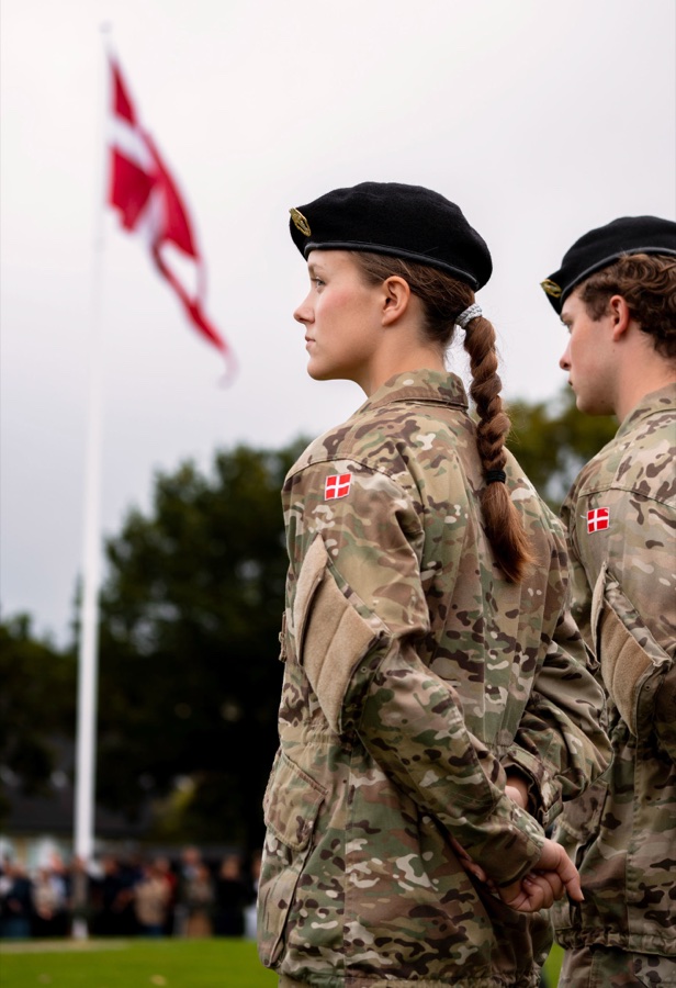 Princess Isabella stands at attention in uniform with a black beret and a plaited braid, the Danish flag flying behind her