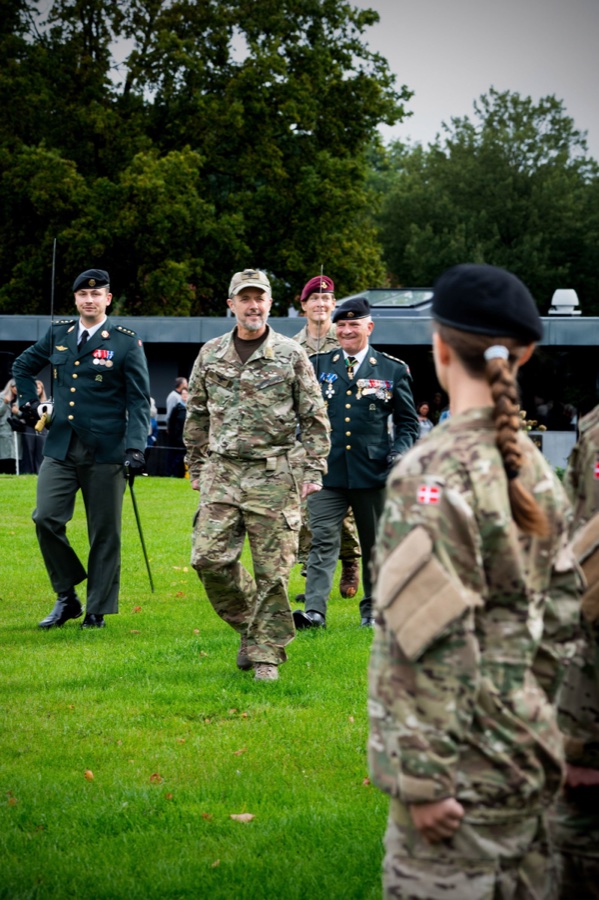 King Frederik in camouflage uniform walks across the grass past the ranks of recruits, with Princess Isabella in the foreground seen from behind