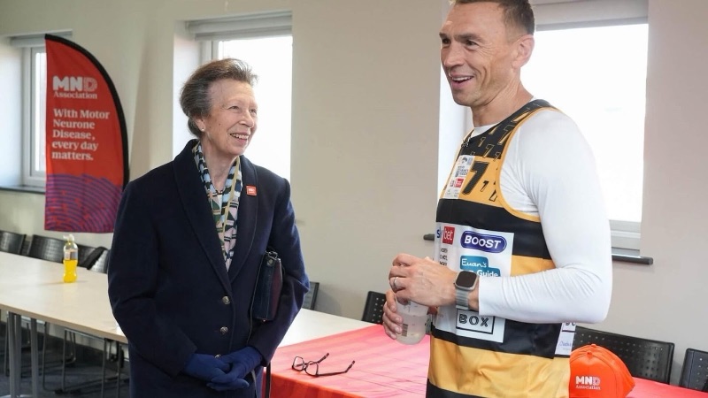 Princess Anne in a dark coat in conversation with Kevin Sinfield in a running vest, in front of a red MND Association banner