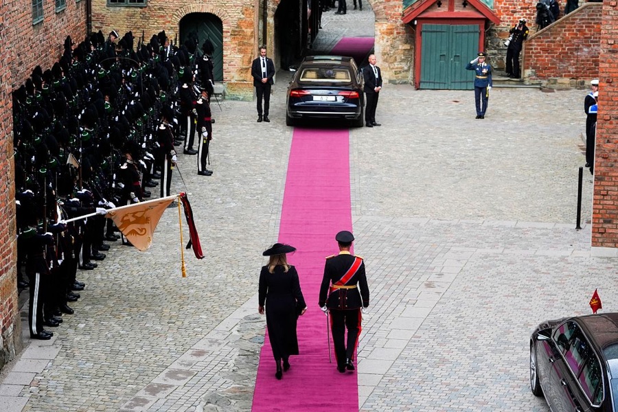 King Haakon in uniform and Queen Mette-Marit in black walk along a pink carpet at Akershus, with a guard of honour drawn up along the wall and the car A 2 at the end