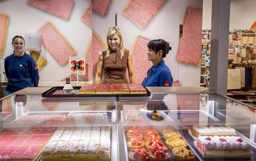 Dronning Máxima står bak en bakerdisk full av kaker og snakker med to ansatte, veggen bak er dekorert med rosa kaker