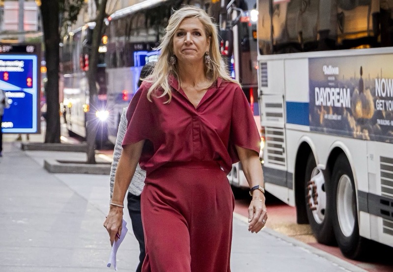 Dronning Máxima går på et fortau i New York om kvelden, med busser og bytrafikk i bakgrunnen