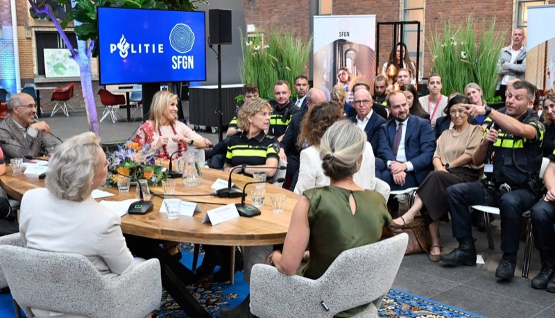 Dronning Máxima sitter ved et rundt bord med mikrofoner, omgitt av politifolk og ledere, foran en skjerm med logoene til politiet og SFGN