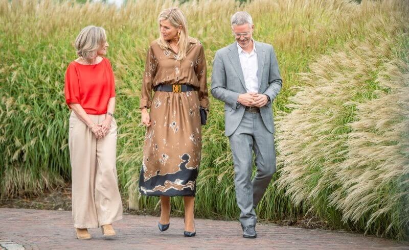 Queen Máxima walks along a path between tall ornamental grasses with a woman in a red blouse and a man in a grey suit