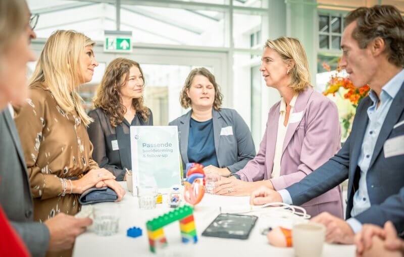 Queen Máxima sits at a table with workshop participants, an anatomical heart model and building blocks on the table