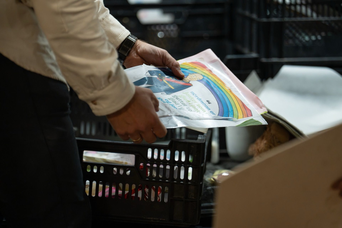 A hand places a child\u0027s drawing of King Harald with a rainbow into a plastic crate at the archive