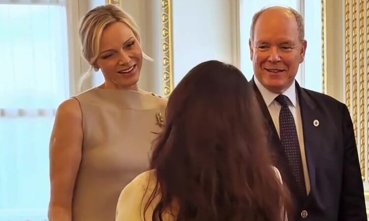 Princess Charlène and Prince Albert smile at a guest standing with her back to the camera