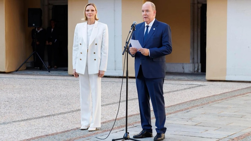 Prince Albert gives an address at a microphone in the palace courtyard, with Princess Charlene standing beside him