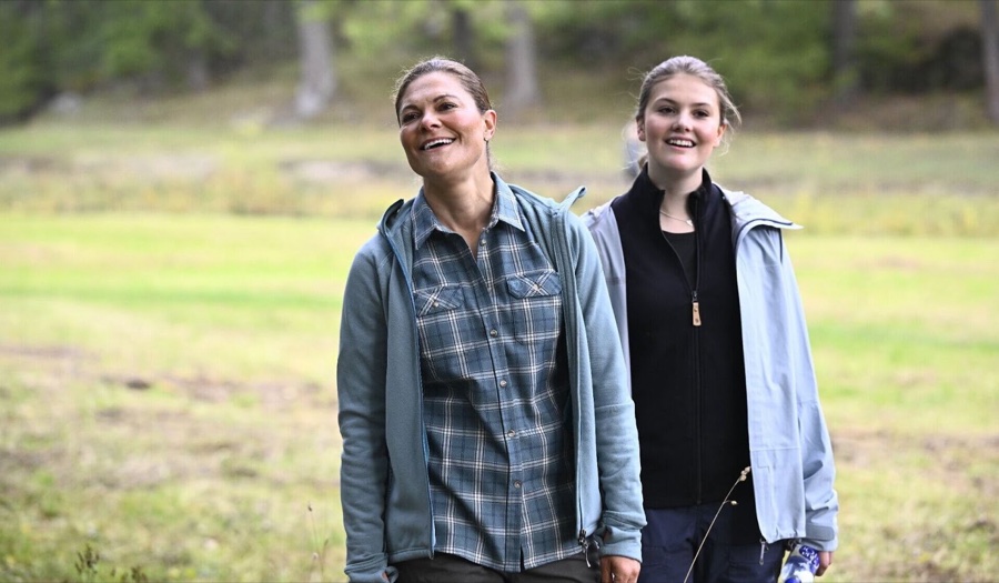 Kronprinsesse Victoria i rutete skjorte og prinsesse Estelle går smilende over en eng med skog i bakgrunnen