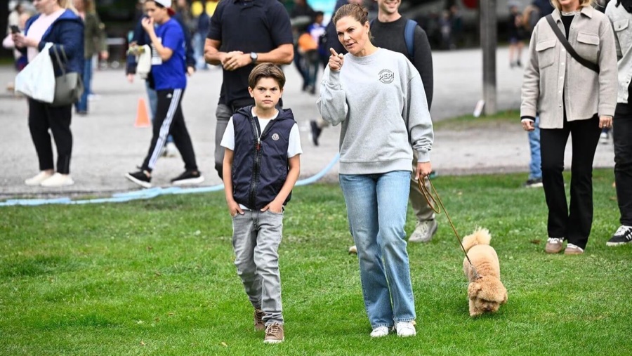 Kronprinsesse Victoria i grå genser med Pep-logo går over gresset sammen med prins Oscar og en liten lys hund i bånd