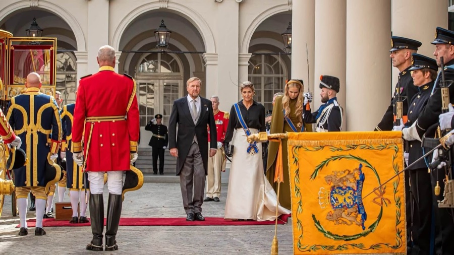 King Willem-Alexander and Queen Máxima walk across a courtyard between guards of honour, with a gilded carriage to the left