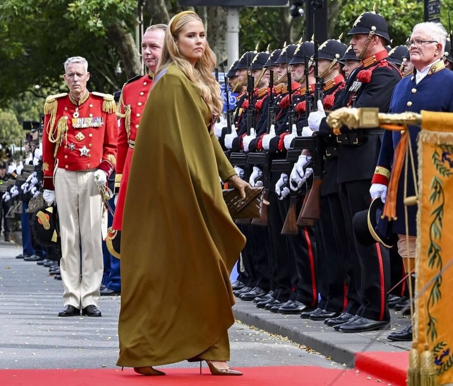 Crown Princess Amalia in a long olive-green cape walks along a line of soldiers on a red carpet