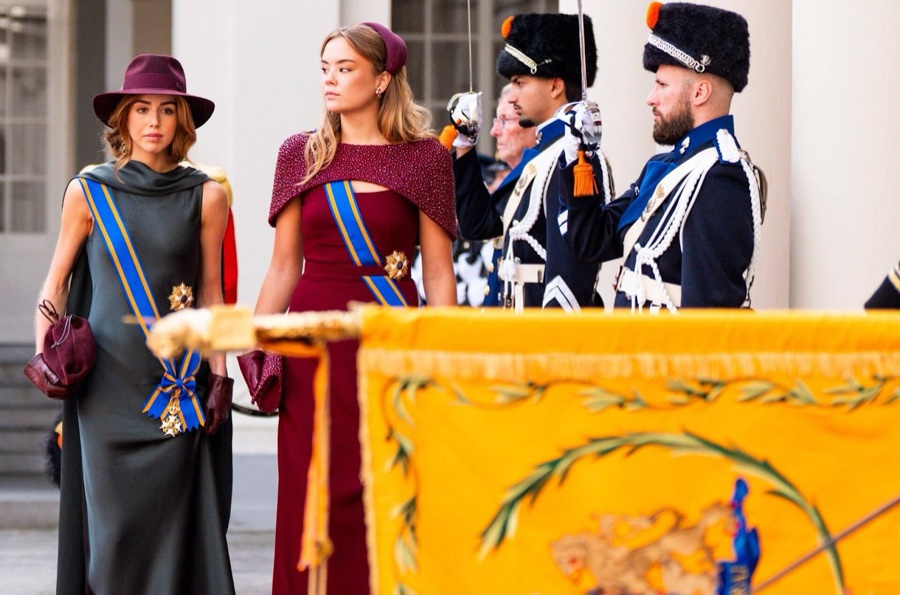 Princess Alexia in a green dress and wide-brimmed hat walks beside Princess Ariane in a ruby-red dress with an embroidered cape